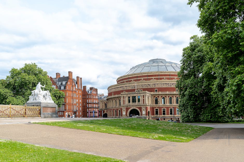 A large, historic theatre with a rounded, domed roof and detailed brickwork features prominently in a cityscape, situated behind a well-maintained grassy area with a wide walkway. To the left side, there is a white stone sculpture with multiple figures and a decorative base, positioned near a metal fence. Tall, multi-story red-brick residential buildings with chimneys and gabled roofs are seen in the background, alongside lush green trees on both sides of the scene. The sky is partly cloudy with patches of blue, and the overall environment appears peaceful and suitable for urban house removals or home relocation activities. This setting is representative of a typical busy city environment with space for loading and unloading furniture, boxes, and relocation equipment, relevant to professional removal services such as Kensington Man and Van.