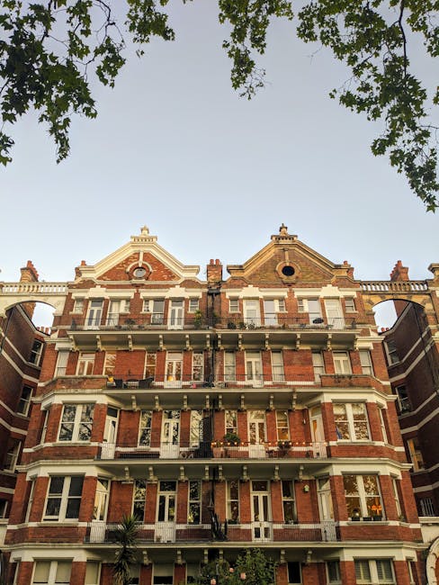 A tall, multi-storey residential building constructed with red brick and cream stone detailing, featuring multiple balconies with black metal railings and large windows. The building has ornate architectural features including decorative gables at the roofline, with some circular window openings on the upper floors. The photo is taken from street level, with a portion of the sidewalk and some greenery, such as a palm tree, visible at the base. Overhanging tree branches frame the top of the image, and the sky is clear with natural daylight illuminating the facade. This scene depicts an urban housing setting suitable for home relocation or furniture transport, as seen in professional removals services like those provided by Kensington Man and Van.