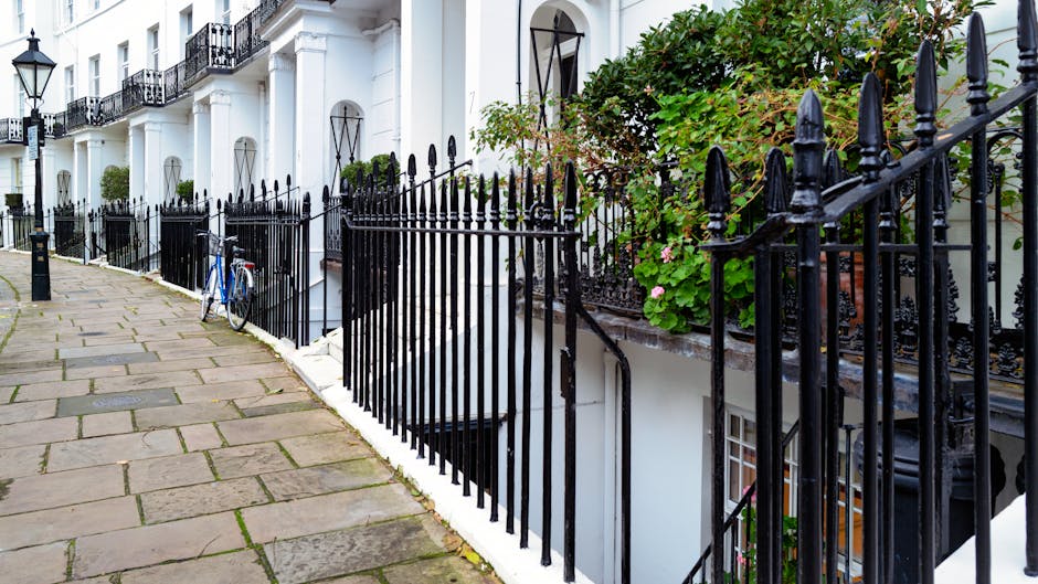 A residential street featuring a white multi-storey building with black wrought-iron railings along the sidewalk. The pavement is made of large, rectangular stone slabs, with some patches of moss or dirt visible between them. A blue bicycle leans against the railing near the building’s entrance, which is accessible via a small staircase with metal handrails. Green leafy plants and flowering bushes grow behind and around the railings, adding greenery to the scene. The building has several windows with black security bars, and the overall setting is well-lit with natural daylight, demonstrating an urban environment suitable for house removals. This image illustrates the typical exterior environment for household relocation planning, as part of professional moving and furniture transport services provided by Kensington Man and Van.