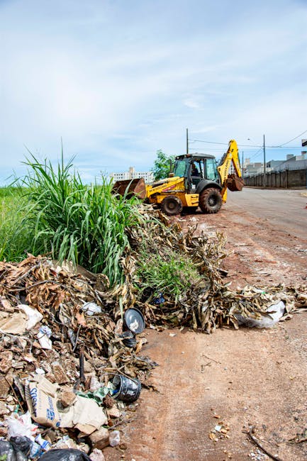 The image shows a large pile of mixed domestic waste, including brown cardboard, crushed plastic bottles, black plastic bags, and various paper materials, situated on a roadside verge. Behind the waste, there is a yellow backhoe loader with a raised front bucket and extended arm, positioned on a dirt and gravel surface adjacent to a paved road. To the left, tall green grass and weeds border the waste pile, while the background features a partly cloudy sky with light blue tones and scattered clouds. In the distance, a high metal fence runs along the edge of the property, with some buildings and utility poles visible further back. The scene illustrates a waste disposal or clearance site, which could relate to bulk waste removal as part of a home relocation or property clearance service. The scene highlights the importance of proper waste management in residential moving or clearance processes, aligning with Kensington Man and Van's removal and disposal services documented on the Kensington bulky waste rules page.