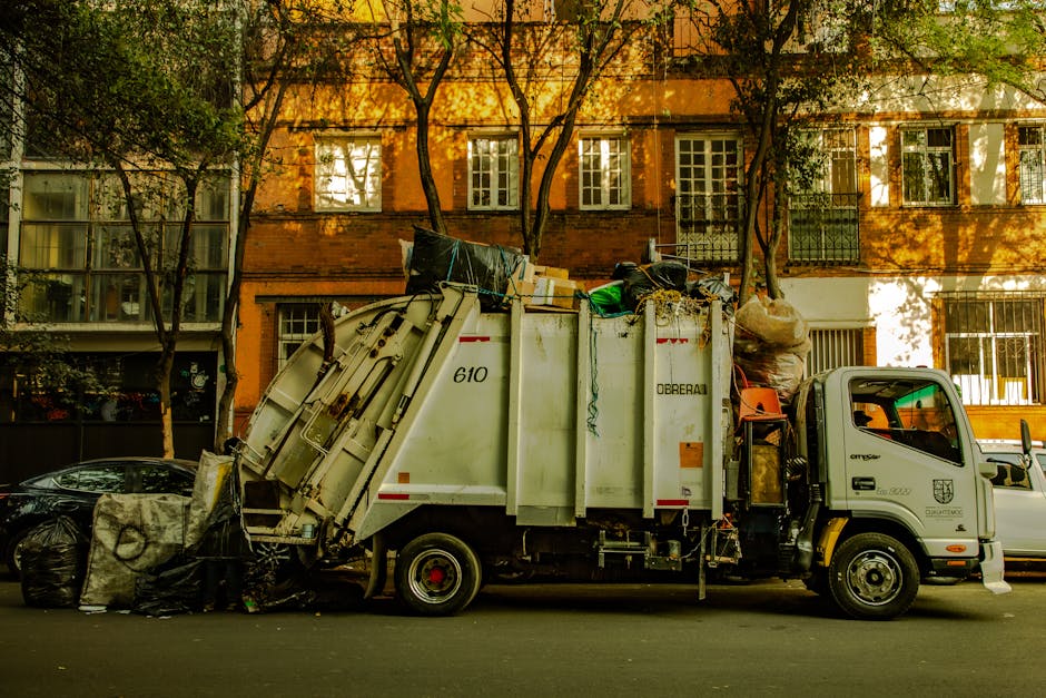 A white waste collection truck with the number 610 and the label 'O'Drebail' on its side is parked on a residential street during daytime. The vehicle is filled with various rubbish, including large plastic bags, cardboard boxes, and discarded household items, indicating a home relocation clean-up or disposal process. The back of the truck is open, with waste materials visibly loaded inside, including fabric, plastic, and cardboard, some stacked and secured with straps. In front of the truck, there are extra trash bags and waste materials placed on the pavement, ready for disposal. The background features a multi-storey red brick building with white-framed windows and balconies, as well as trees casting shadows on the facade. The scene suggests an organized collection of household waste, potentially related to house removals, and is indicative of the kind of waste management services provided by Kensington Man and Van or similar removals companies in the area.