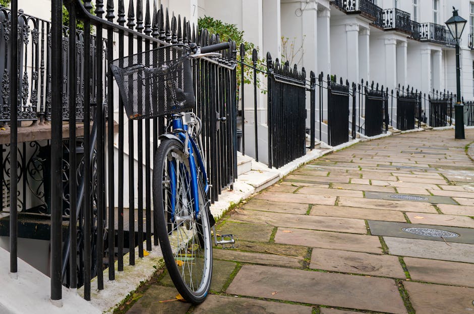 A blue bicycle with a front metal basket leaning against a black wrought iron fence outside a row of white townhouse buildings on a curved, stone-paved sidewalk. The bicycles are situated next to a white stone staircase leading to the residential entrance, which is partially visible behind the fence. The area is well-lit, with sunlight casting shadows on the pavement. The image depicts a typical London street scene, with the fence separating the sidewalk from the building steps. This setting relates to house removals or property relocation services provided by Kensington Man and Van, showcasing the typical urban environment where furniture and household items might be transported through narrow and accessible pathways during a home relocation or moving process.