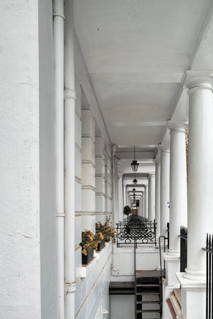 A narrow exterior street corridor with white-painted stone or concrete buildings featuring classic architectural details, including tall columns, pilasters, and decorative lamp posts suspended from the ceiling. Along the side of the buildings, there are flower boxes with small, dried plants. The corridor is partially shaded with natural light, and the image captures a sequence of black metal railings and steps leading downwards, suggesting an entrance or basement access. This setting is part of a residential area consistent with a typical London neighbourhood like Kensington. The image reflects a property secured with external access points suitable for home relocations or furniture transport, as coordinated by a professional removals company such as Kensington Man and Van. The overall scene emphasizes the building's historic charm and the logistical considerations of moving large or heavy items through confined, hard-to-access streets during a home removal or packing and moving process.