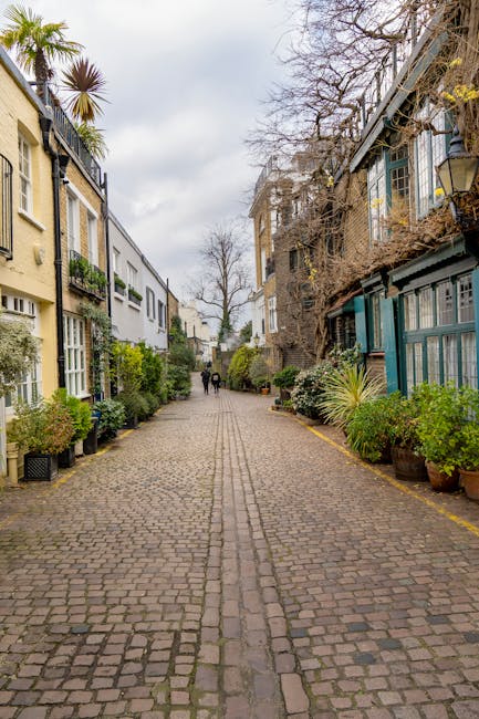 A narrow cobbled street in a residential area during daytime, flanked by multi-storey buildings with a mix of modern and traditional architecture, some with balconies and window boxes full of plants. On the right side, there is a blue-painted building with a metal staircase and potted plants along the pavement. On the left, a pale yellow building with large windows and greenery outside. In the distance, a couple of pedestrians are walking along the street. The overcast sky overhead casts diffuse natural light. The scene depicts a quiet urban environment suitable for home relocation or furniture transport services, with visible paving stones and urban foliage. Kensington Man and Van provides removals services in such narrow, hard-to-access streets, assisting with packing, loading, and transport processes in busy city environments.