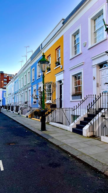 The image depicts a row of residential terraced houses on a quiet street in Kensington, London, painted in pastel shades including light blue, yellow, and lavender. The houses feature white-framed sash windows, some with flower boxes on the window sills, and front doors accessed by small staircases with black metal railings. A traditional black lamppost stands on the pavement near the houses. The street is lined with a paved sidewalk and a narrow asphalt road without visible traffic. In the context of house removals, the scene suggests the exterior environment where furniture and boxes might be loaded into a van, with Kensington Man and Van professional movers preparing to facilitate home relocation or furniture transport. The natural lighting indicates daytime, with clear skies overhead. This setting exemplifies typical London residential streets prepared for packing, loading, and moving services, emphasizing the importance of careful handling of goods during the loading process in a professional removal operation.