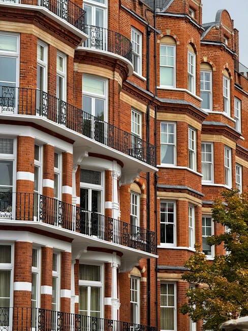 The image depicts a row of residential terraced houses on a quiet street in Kensington, London, painted in pastel shades including light blue, yellow, and lavender. The houses feature white-framed sash windows, some with flower boxes on the window sills, and front doors accessed by small staircases with black metal railings. A traditional black lamppost stands on the pavement near the houses. The street is lined with a paved sidewalk and a narrow asphalt road without visible traffic. In the context of house removals, the scene suggests the exterior environment where furniture and boxes might be loaded into a van, with Kensington Man and Van professional movers preparing to facilitate home relocation or furniture transport. The natural lighting indicates daytime, with clear skies overhead. This setting exemplifies typical London residential streets prepared for packing, loading, and moving services, emphasizing the importance of careful handling of goods during the loading process in a professional removal operation.