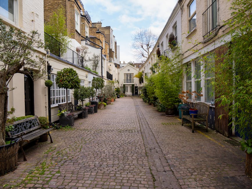 A narrow residential street paved with cobblestones, lined with white-painted terraced houses featuring large windows and balconies, some with black metal railings and potted plants. Along the street,