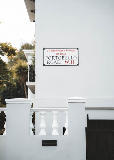 Close-up of a white residential building wall with a street sign reading 'The Royal Borough of Kensington and Chelsea, Portobello Road, W.11' mounted on it. Below the sign, a small black mailbox is embedded in the wall. The lower part of the building features a decorative white balustrade with vertical balusters, partially visible in front of a black gate or fence. The scene is well-lit with natural daylight, with some greenery, including trees and bushes, visible in the background. This setting depicts a typical house frontage suitable for visual reference in content related to house removals or home relocation services, highlighting aspects such as building exterior and street signage within the Kensington area.