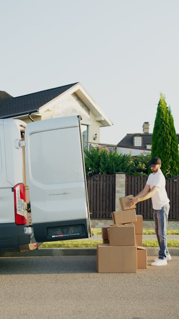 A man wearing a white t-shirt and jeans is standing outside on a residential street during daytime, preparing cardboard boxes for loading into a white cargo van operated by Kensington Man and Van. The