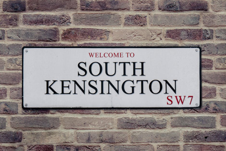 A rectangular white metal sign mounted on a red brick wall displaying the text 'WELCOME TO SOUTH KENSINGTON SW7' with 'South Kensington' written in large black capital letters, 'WELCOME TO' and 'SW7' in smaller red fonts, indicating a street or area in London. The brick wall has a textured surface with visible mortar joints and a slightly weathered appearance. The sign is positioned centrally on the wall, which provides a stable backdrop for depicting a typical London residential or commercial area. This image may be relevant to house removals or local property services, highlighting the environment where packing, furniture transport, and home relocation activities by Kensington Man and Van could typically occur.